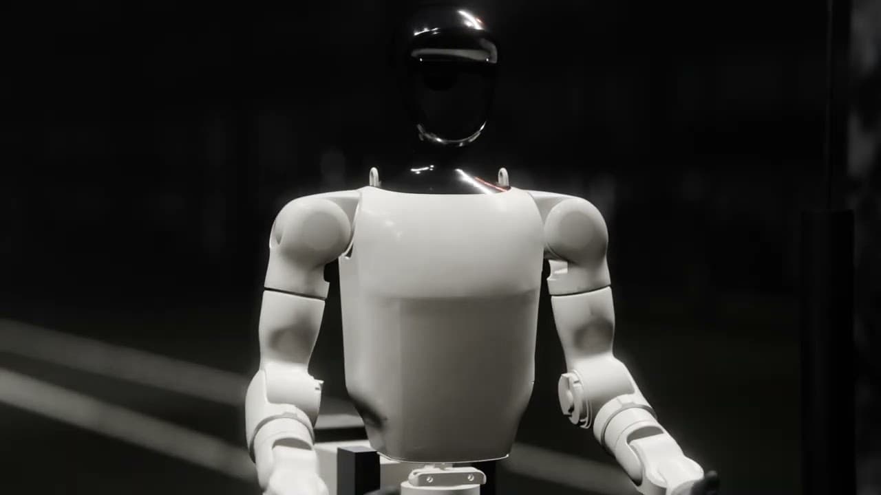 White humanoid robot with a glossy black visor, standing alone in a dark factory hall.