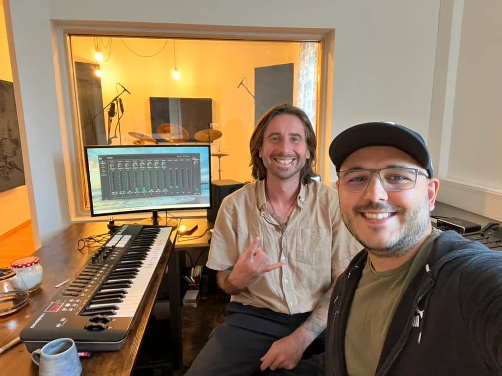 Two people smiling in a recording studio beside a keyboard and mixing screen, with a drum kit visible through the window.